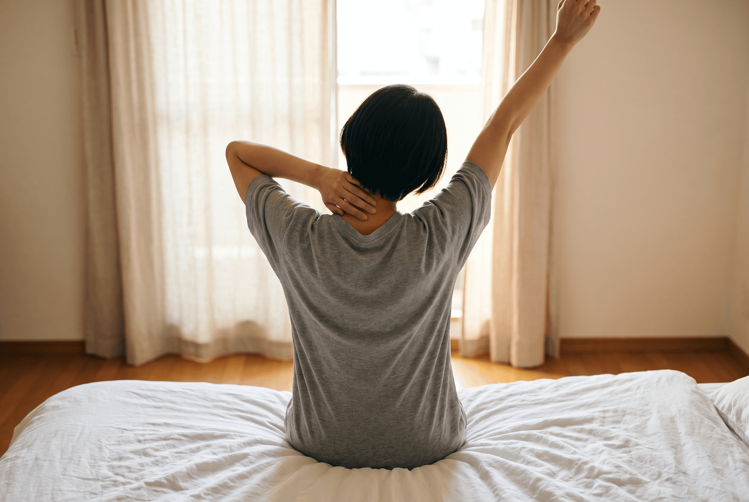 A person seen from behind, sitting on a white bed and stretching one arm overhead towards a bright window.