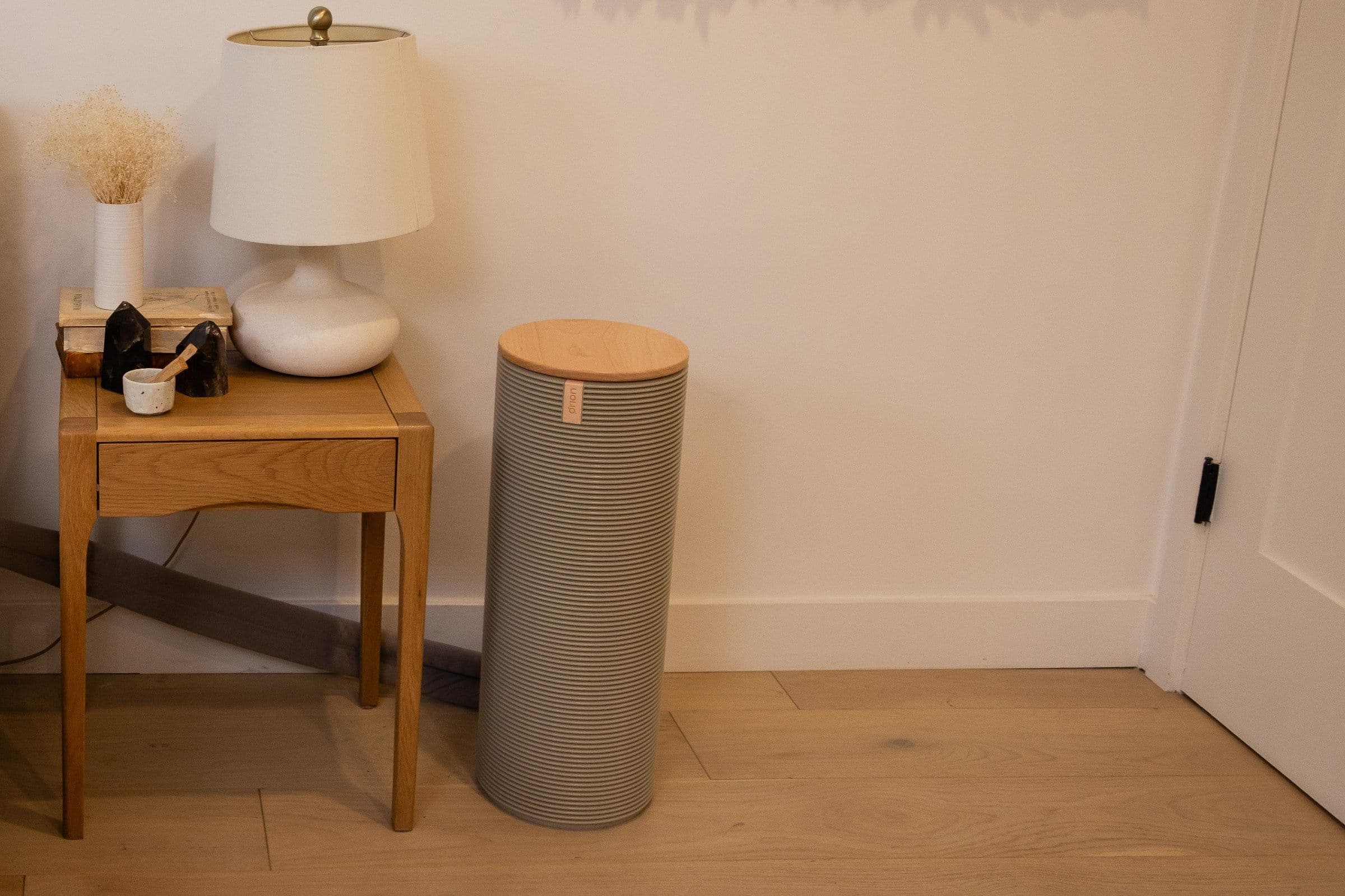 The Orion Control Tower, a ribbed grey cylinder with a wooden lid, standing on the floor beside an oak nightstand with a white lamp.