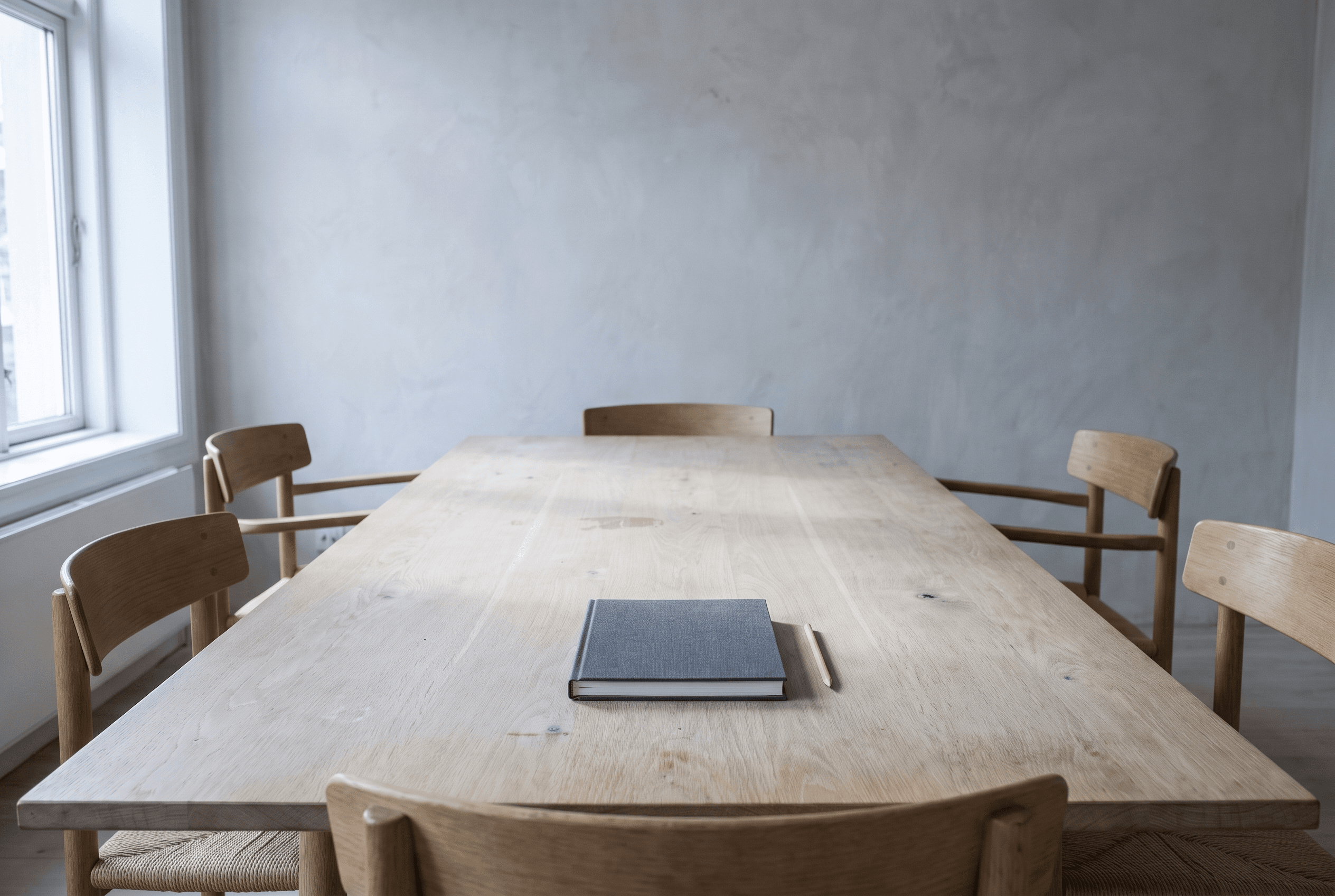 Empty pale oak meeting table with a closed grey notebook and pencil, wooden chairs around it, window light