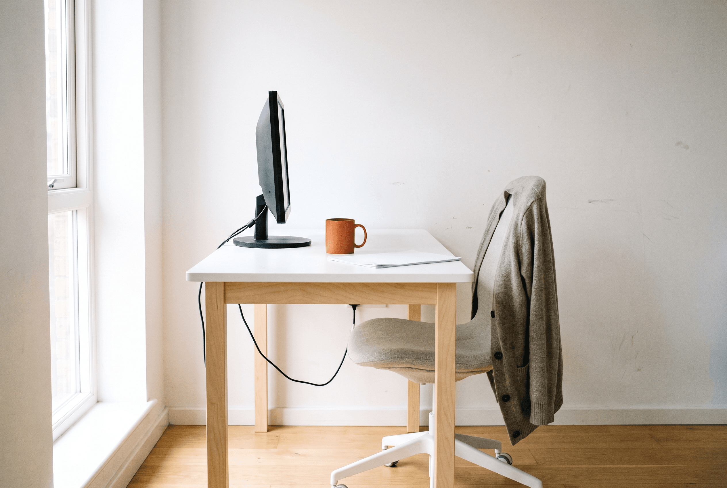 Side view of a white desk by a window with a low monitor, orange mug and a cardigan-draped chair