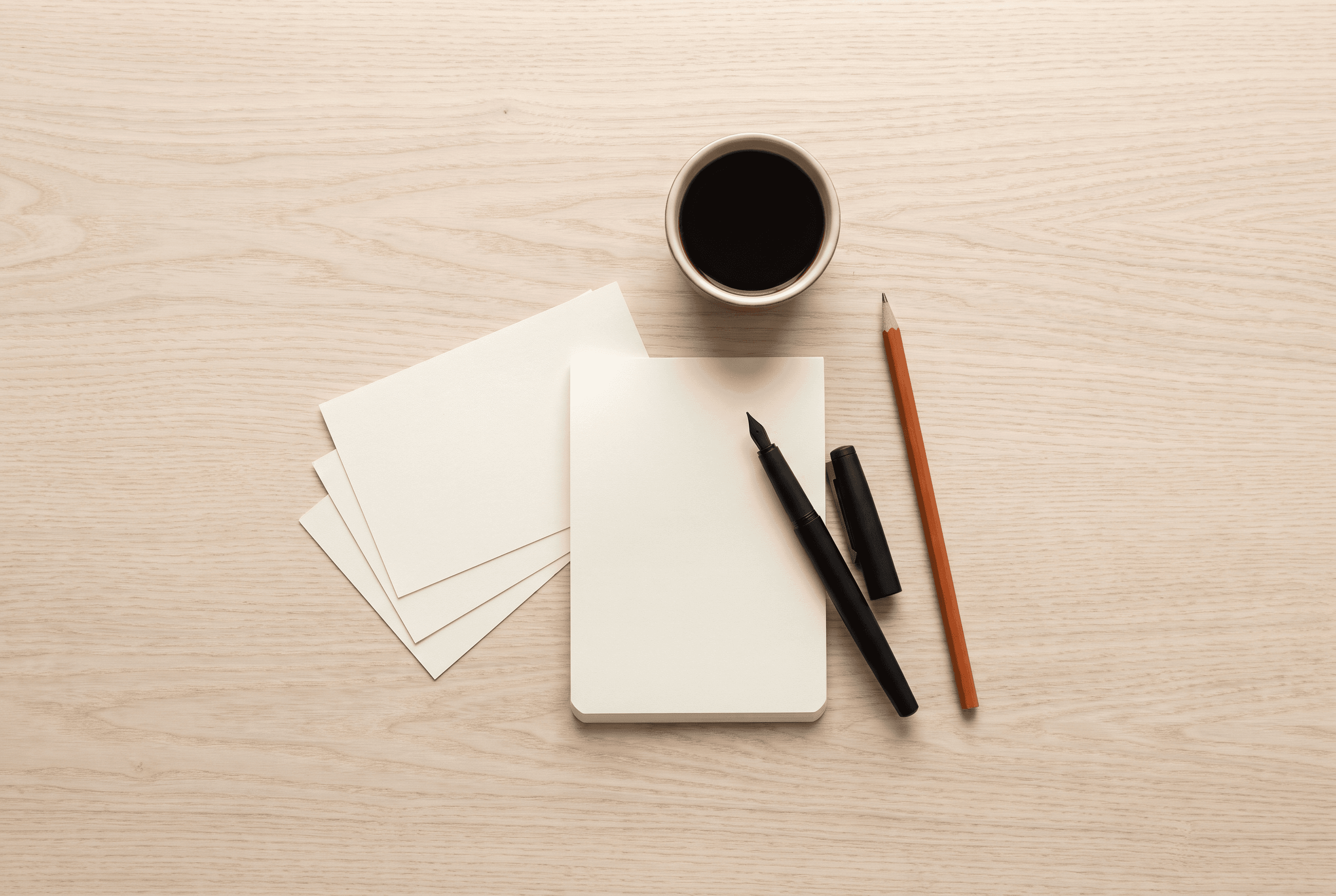 Overhead view of a blank notepad, blank index cards, a black fountain pen, a pencil and a cup of coffee